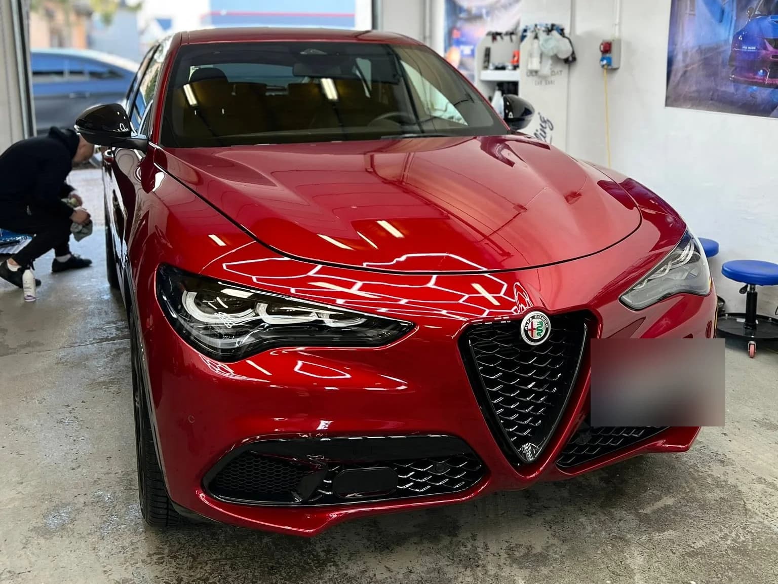 Czerwona Alfa Romeo Stelvio w hali studia, obok pracownik przy elementach nadwozia — lakier lustrzanie odbija oświetlenie.