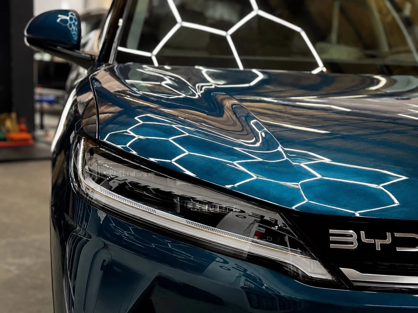 Zbliżenie maski turkusowego BYD Atto 3 po nałożeniu powłoki — w lakierze odbijają się plastry lamp sufitowych.