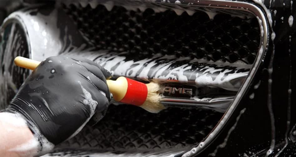 Dłoń w rękawicy czyści pędzelkiem detailingowym grill Mercedesa AMG pokryty pianą