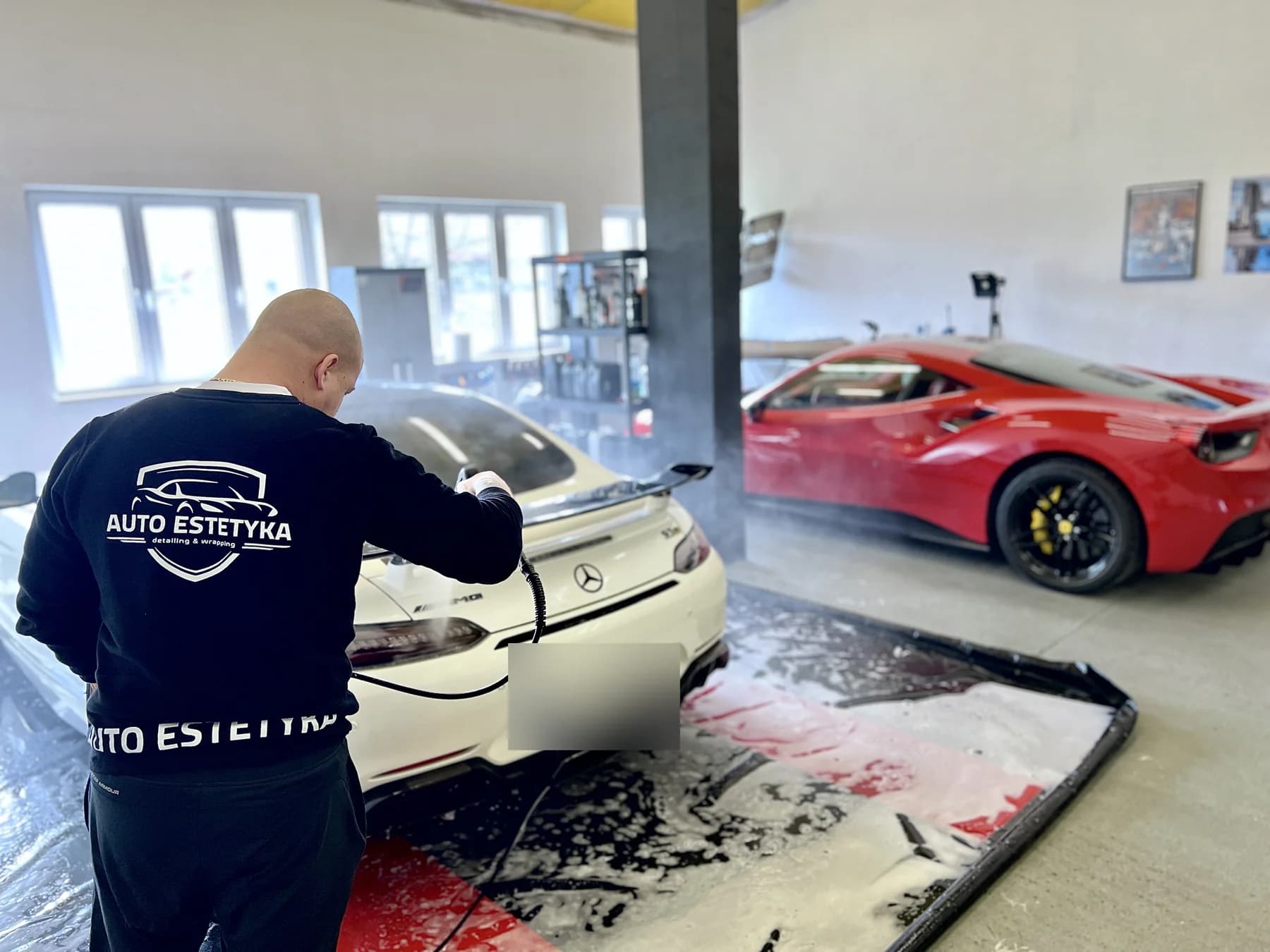 Pracownik Auto Estetyki w firmowej bluzie myje białego Mercedesa-AMG GT na macie do mycia; w tle czerwone Ferrari.