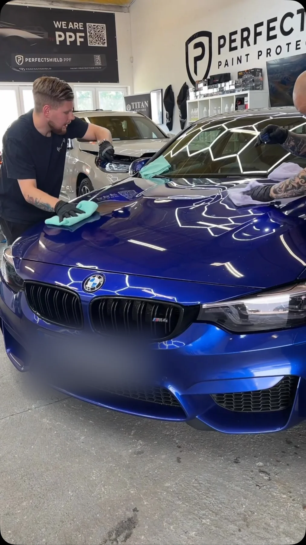 Dwóch detailerów pracuje nad korektą lakieru niebieskiego BMW M3 — na masce widoczne odbicia lamp o kształcie plastrów miodu.