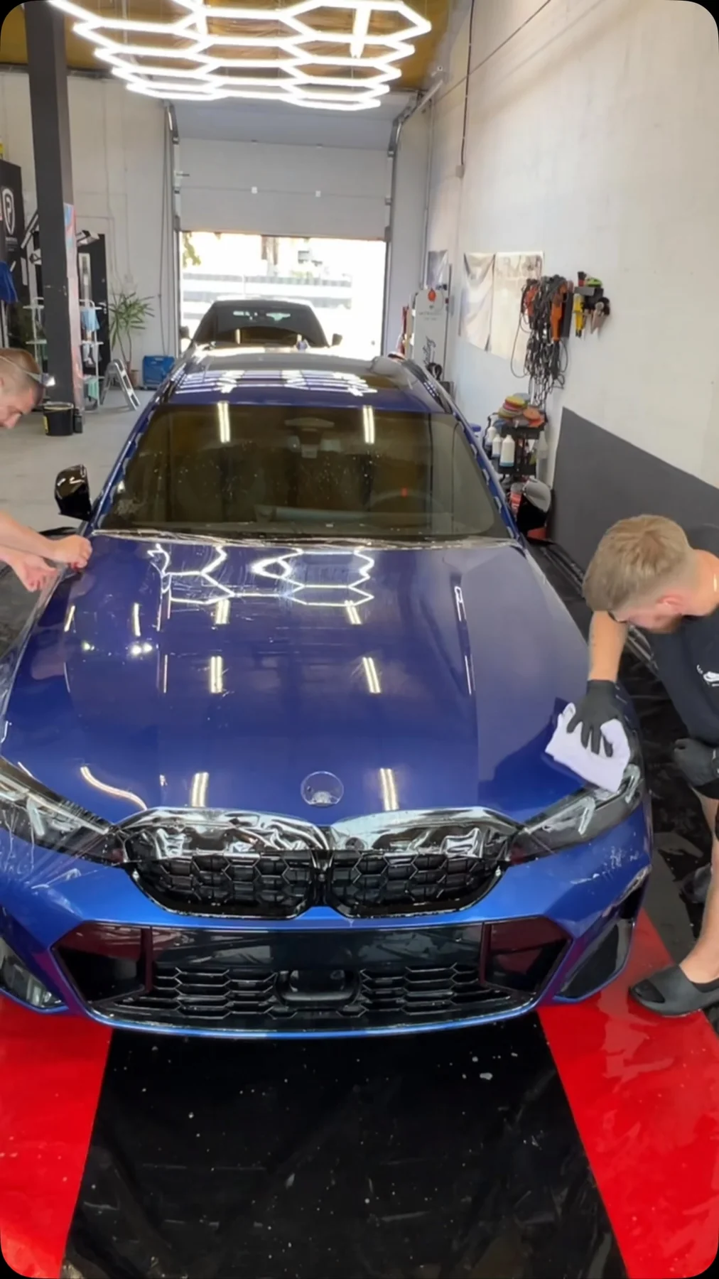 Niebieskie BMW M3 w hali studia pod lampami o kształcie plastrów miodu, dwóch detailerów pracuje przy przodzie auta.