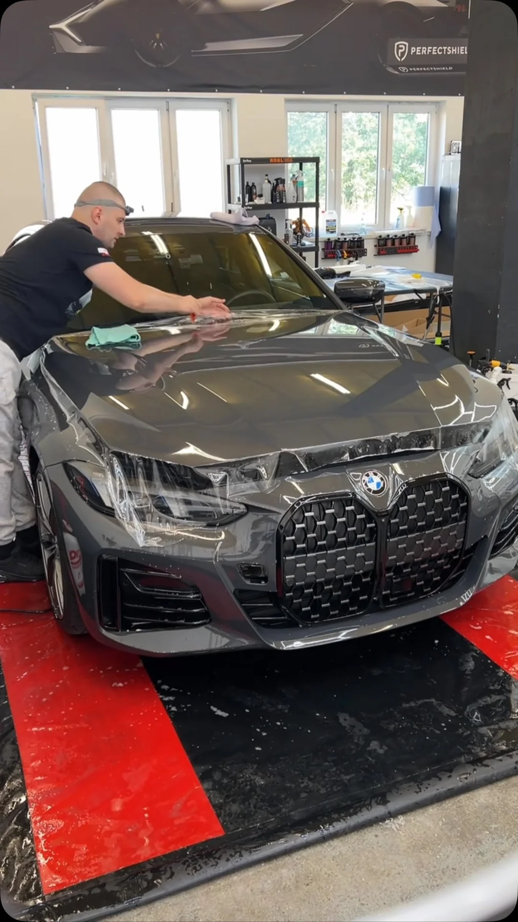 Detailer przeciera maskę szarego BMW M4 przed założeniem folii — auto stoi na czerwonej macie w hali studia.
