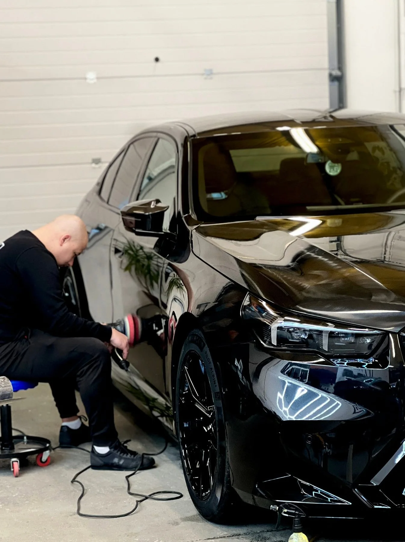 Detailer aplikuje folię ochronną PPF na przednim zderzaku czarnego BMW, pracując na niskim siedzisku warsztatowym.