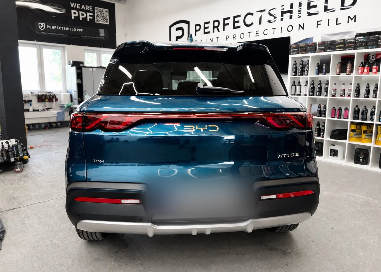 Tył turkusowego BYD Atto 3 w hali studia po nałożeniu powłoki ceramicznej, w tle ekspozycja folii PerfectShield.
